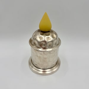 Könnte beinhalten: Ein silberfarbener Behälter mit gehämmerten Texturen und einem gelben, tropfenförmigen Aufsatz. Der Behälter hat einen runden Sockel und einen Deckel. Das Design ist schlicht und elegant, geeignet für die Aufbewahrung kleiner Gegenstände. Maße in Zentimetern.