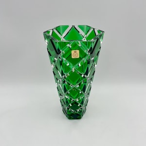 May include: A striking, emerald green glass vase featuring a geometric diamond pattern. The vase has a wide opening and a faceted design, creating a play of light and shadow. A small, gold-toned sticker is affixed to the front.