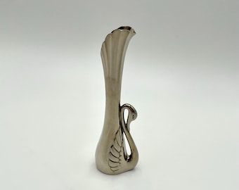 May include: A small, silver-colored vase with a swan design. The vase has a fluted, flared top and a swan's neck and wing forming the body. The swan's wing is detailed with etched lines. The vase is approximately 6 inches tall.