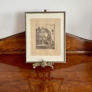 Pode incluir: Uma gravura emoldurada a preto e branco de um edifício com um candeeiro de rua. A obra de arte é exibida numa tela decorativa de latão. A moldura é prateada e a obra de arte está sobre um fundo creme.