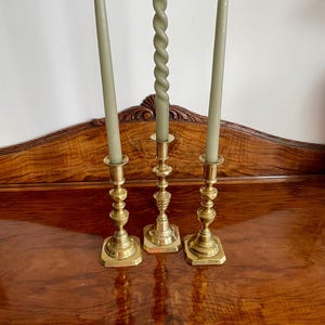 Può includere: Tre candelabri in ottone con candele verde salvia. Due candele dritte affiancano una candela attorcigliata. I candelabri hanno una base quadrata e un design dettagliato a più livelli. Le candele sono spente e poggiano su una superficie di legno scuro.