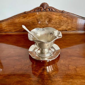 May include: A silver gravy boat with a scalloped rim, resting on a matching saucer. A small spoon rests inside the boat. The set is made of a shiny, reflective metal, likely silver or a silver-toned alloy. The gravy boat and saucer are placed on a polished wooden surface.