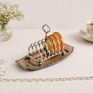 May include: A silver-toned toast rack holding several slices of golden-brown toast. The rack has a decorative handle and sits on a matching rectangular tray. The setting includes a vase of white flowers and a cup of tea.