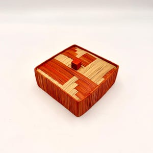 May include: A square lidded box crafted from woven fibers in alternating shades of orange and cream. The lid features a geometric pattern of diagonal strips and is topped with a small, solid orange cube handle centered on the top surface.