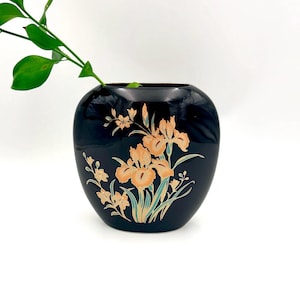 Può includere: Un vaso in ceramica nera con finitura lucida, decorato con un motivo floreale con iris color pesca e fiori più piccoli color crema. Foglie e steli verdi completano il design. Il vaso contiene una pianta verde.