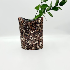 May include: A decorative vase with a unique pattern and a black rim. The vase is brown with gold and white abstract designs. Green leaves and stems are placed inside the vase. The vase is on a white surface.