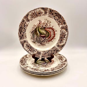 May include: A stack of three round ceramic plates featuring a central illustration of a turkey with green, brown, and red plumage. The rim is decorated with a brown floral and bird pattern on a cream background, with scalloped edges.