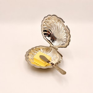 Op de afbeelding: Een zilverkleurige, schelpvormige botervloot met een scharnierend deksel. Binnenin ligt een geel, rechthoekig stukje boter met een klein botermes. De schaal heeft een decoratieve, geschulpte rand en is open.