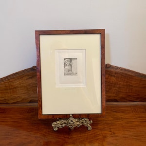 May include: A framed black and white print of a person in a hat, displayed on a decorative brass stand. The frame is a dark wood with a light cream mat. The stand is ornate with a gold finish. The print is displayed on a wooden surface.