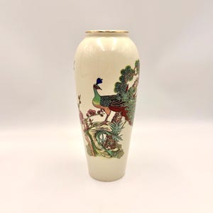 Może przedstawiać: Kremowy wazon ceramiczny ze szczegółową ilustracją pawia stojącego na skale, otoczonego gałęziami sosny i elementami kwiatowymi. Wazon ma zwężający się cylindryczny kształt i cienką złotą obwódkę przy górnym otworze.