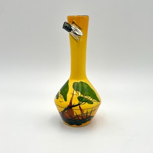 Op de afbeelding: Een gele keramische vaas met een slanke hals en een bolvormige basis. De vaas heeft een handgeschilderde scène van een schip en bomen. Een decoratieve zilverkleurige bij is bevestigd aan de hals van de vaas.
