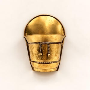 Op de afbeelding: Een aan de muur gemonteerde koperen plantenbak met een halfronde achterplaat en een taps toelopende emmer met een gehamerde textuur. De emmer is versierd met twee horizontale touwachtige banden en een centraal decoratief touwdetail met twee kleine bolvormige knoppen.