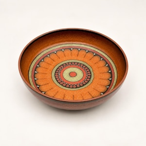 May include: A round ceramic bowl featuring a vibrant orange interior with a central circular pattern. The design includes concentric rings in shades of muted green, black, and red, surrounded by a stylized petal motif in orange and black.