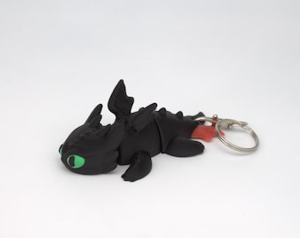 May include: A black dragon keychain with green eyes and a red tongue. The dragon has detailed wings and a silver keyring attached. The dragon is lying down on a white surface.