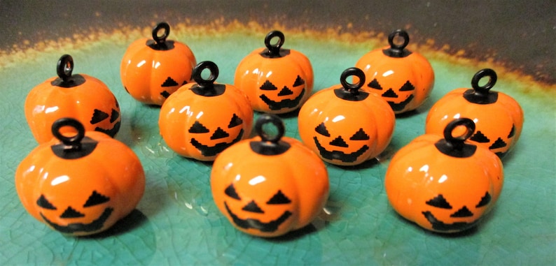 May include: Ten orange pumpkin charms with black jack-o'-lantern faces and black metal loops. The charms are on a teal surface.