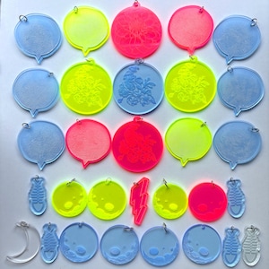 May include: A collection of colorful acrylic charms in various shapes, including speech bubbles, circles, moons, fish, and a lightning bolt. The charms are in shades of blue, yellow, pink, and clear, with some featuring etched floral designs.