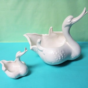 May include: Two white ceramic swan-shaped planters. The larger swan has a long neck and a wide, open body. The smaller swan has a similar design. Both are on a turquoise surface, with a green background.