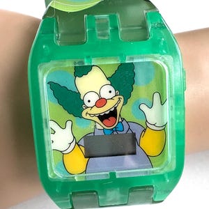May include: A green digital watch with a cartoon Krusty the Clown image. The watch has a clear face and a green plastic strap. The clown has green hair, a red nose, and a wide smile. The watch is on a wrist.