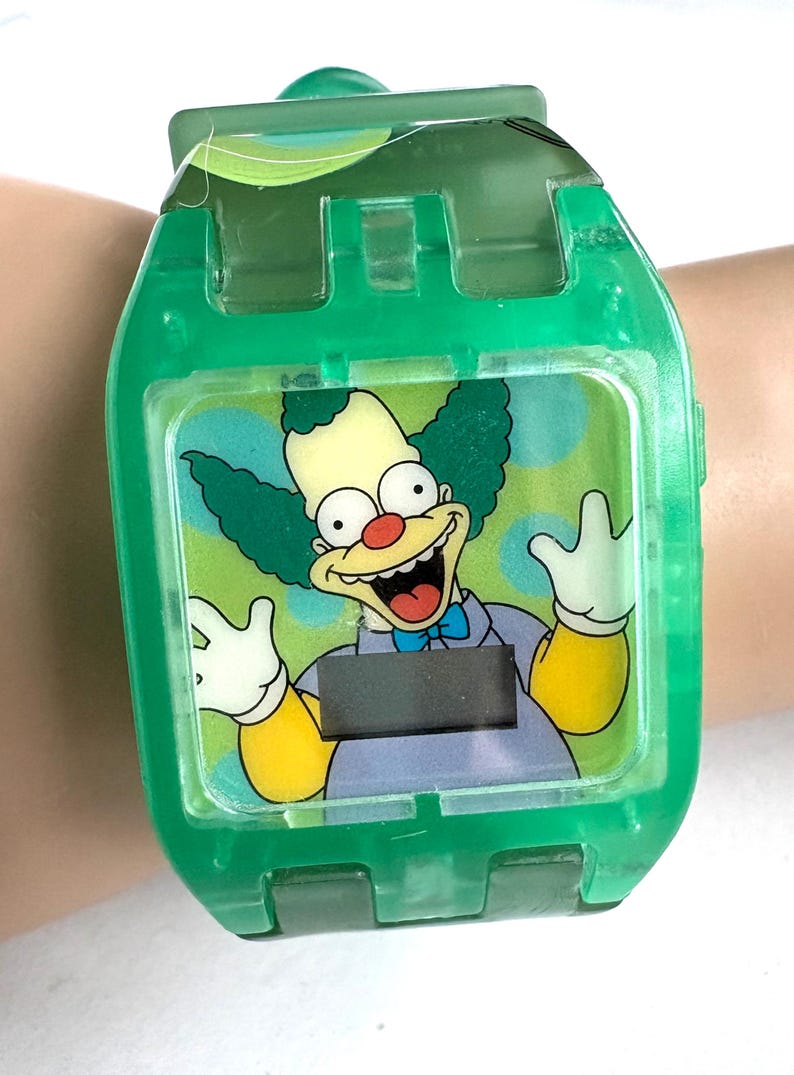 May include: A green digital watch with a cartoon Krusty the Clown image. The watch has a clear face and a green plastic strap. The clown has green hair, a red nose, and a wide smile. The watch is on a wrist.
