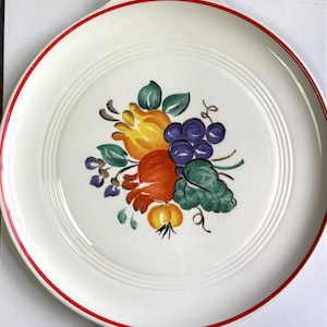 May include: A white ceramic serving platter with a red rim and handles. The center features a colorful illustration of fruit, including grapes, oranges, and lemons. The design is surrounded by concentric circles.