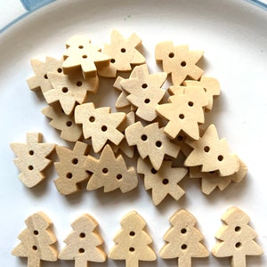 May include: A collection of small, light-colored wooden Christmas tree-shaped buttons. Each button has two holes for sewing. The buttons are scattered on a white plate with a blue rim, perfect for holiday crafts.