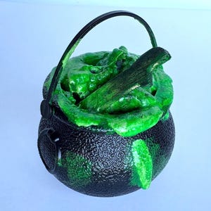 May include: A black plastic cauldron with a textured surface and a black handle. The interior is filled with a bright green, bubbly, viscous substance that overflows the rim. A dark, weathered wooden stick is partially submerged in the green mixture.