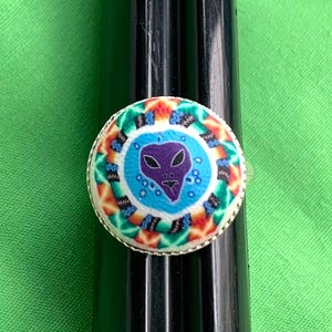 May include: A silver ring with a round, colorful design featuring a purple alien face on a blue background. The ring has a detailed, patterned border with orange, green, and blue elements, set against a green backdrop.