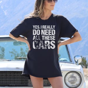 May include: A black short-sleeved t-shirt featuring white distressed-style text that reads, "YES, I REALLY DO NEED ALL THESE CARS." The shirt has a crew neckline and a relaxed fit, with the graphic centered prominently on the chest area.
