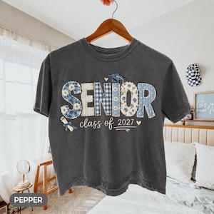 May include: A charcoal gray short-sleeve t-shirt featuring the word SENIOR in large, patterned block letters with blue floral, plaid, and polka dot designs. A graduation cap sits atop the letters, with the text class of 2027 and a diploma graphic.