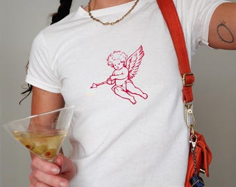 Puede incluir: Camiseta blanca con una ilustración roja de un querubín con arco y flecha. La camiseta se combina con un bolso rojo y un collar dorado. Una copa de martini con aceitunas se sostiene en la mano.