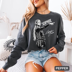 May include: A charcoal gray long-sleeve crewneck sweatshirt featuring a white graphic print of a basketball player pointing, with the name SOPHIE printed above and a signature below. The garment has a relaxed fit and a vintage-washed appearance.