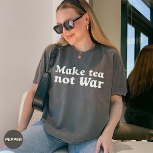 May include: A gray t-shirt with the text "Make tea not War" in white. The shirt is short-sleeved and has a crew neck. The person is wearing sunglasses and a black headband.