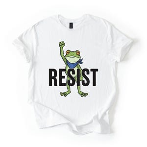 May include: White t-shirt with a cartoon frog design. The frog, wearing a blue bandana, has a raised fist. The word "RESIST" is printed in black below the frog. The frog is green with yellow eyes.