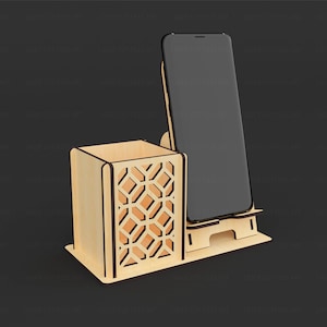 May include: A light-colored wooden desk organizer featuring a square pen holder with a geometric cutout pattern on the side and an integrated upright stand designed to hold a smartphone in a vertical position on a flat rectangular base.