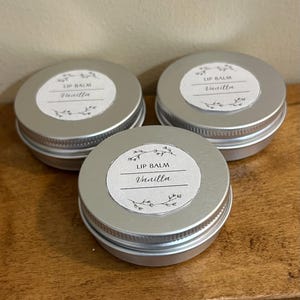 Puede incluir: Tres latas de bálsamo labial plateadas con etiquetas blancas que dicen "Lip Balm Vanilla".