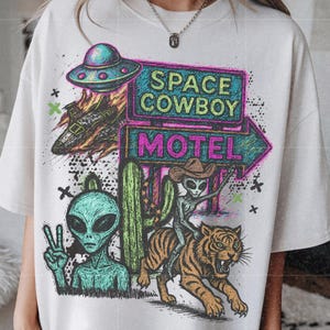 May include: Cream-colored t-shirt featuring a colorful graphic with an alien theme. The design includes a UFO, a cactus, and an alien riding a tiger. Text on the shirt reads "SPACE COWBOY MOTEL."