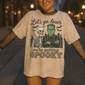 K&ouml;nnte beinhalten: Beiges T-Shirt mit einem Halloween-Motiv. Das Motiv zeigt ein Skelett mit einem Getr&auml;nk, Frankensteins Monster, Flederm&auml;use und den Text "Let's go, loser, we're getting spooky."