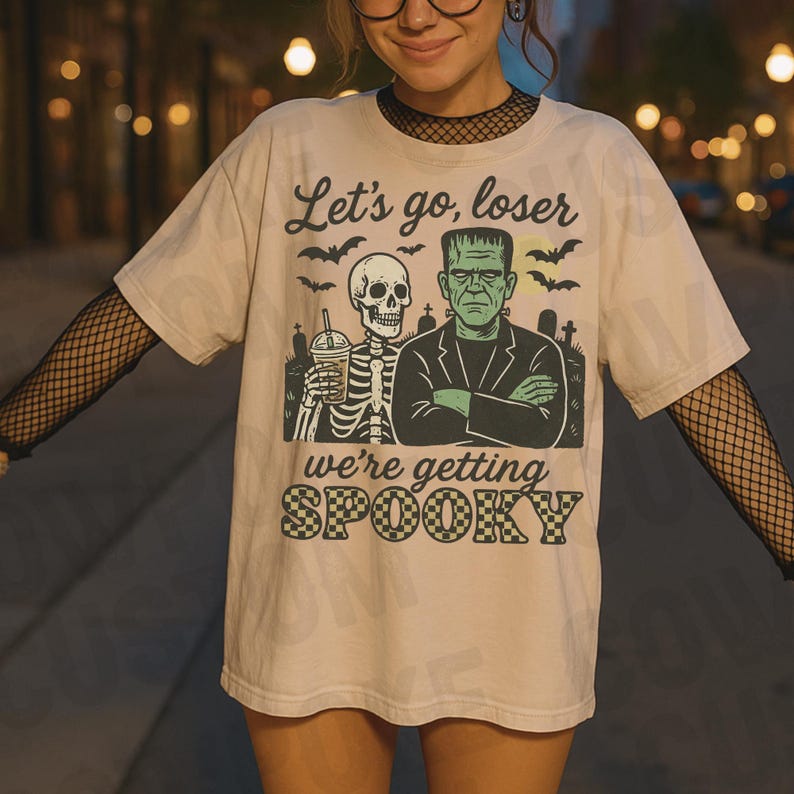 K&ouml;nnte beinhalten: Beiges T-Shirt mit einem Halloween-Motiv. Das Motiv zeigt ein Skelett mit einem Getr&auml;nk, Frankensteins Monster, Flederm&auml;use und den Text "Let's go, loser, we're getting spooky."