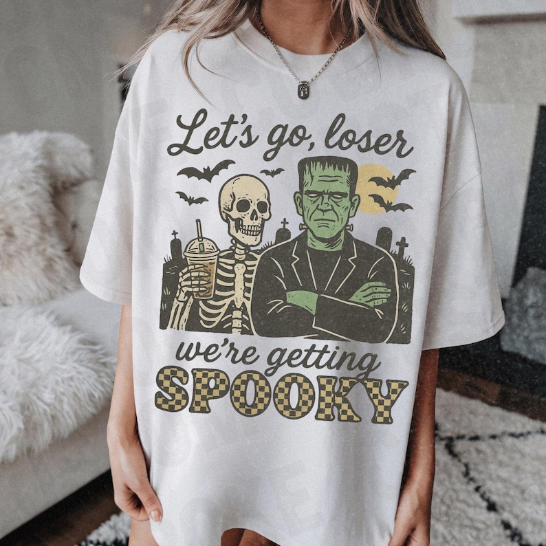 K&ouml;nnte beinhalten: Cremefarbenes T-Shirt mit einem Halloween-Motiv. Das Design zeigt ein Skelett mit einem Getr&auml;nk, Frankensteins Monster, Flederm&auml;use und den Text "Let's go, loser, we're getting SPOOKY."
