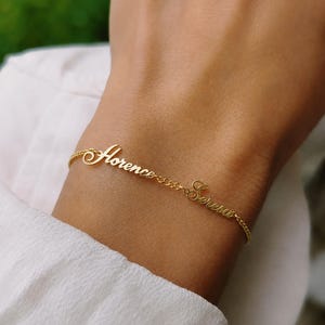 Puede incluir: Un brazalete dorado con los nombres "Florence" y "Serena" escritos en una delicada caligrafía. El brazalete está unido a una fina cadena. La imagen muestra el brazalete en una muñeca, con un fondo borroso.