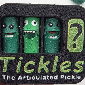 May include: A black rectangular container with three green articulated pickle toys inside, each with a unique facial expression. The container has the word "Tickles" in green and yellow, with "The Articulated Pickle" below. A green question mark is on the right.