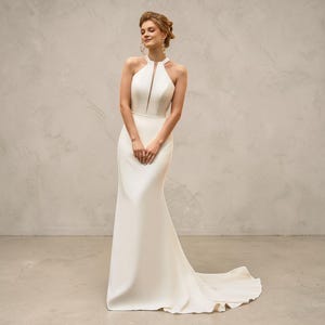 May include: A floor-length white gown featuring a halter neckline with a central vertical cutout, a fitted bodice, and a long skirt that flares slightly at the hem with a train. The dress has a smooth, solid finish and a clean, structured silhouette.