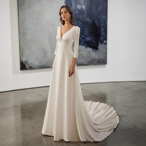 Könnte beinhalten: Ein weißes Brautkleid mit V-Ausschnitt und Dreiviertelärmeln. Das Kleid hat ein figurbetontes Oberteil und einen fließenden Rock, der in eine Schleppe übergeht. Der Stoff ist glatt und elegant. Das Logo "Versailles" ist oben links zu sehen.