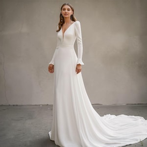 May include: A white bridal gown with a deep V-neck and long sleeves. The dress has a fitted bodice, a flowing skirt, and a long train. The sleeves have a frilled detail at the wrist. The dress is from the "Versailles" collection.