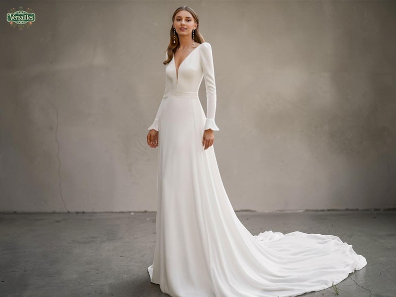 May include: A white bridal gown with a deep V-neck and long sleeves. The dress has a fitted bodice, a flowing skirt, and a long train. The sleeves have a frilled detail at the wrist. The dress is from the "Versailles" collection.