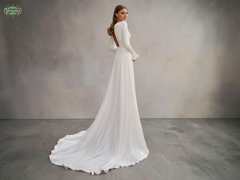 May include: A white wedding dress with a long train and sleeves. The dress has a high neckline and a fitted bodice that flows into a full skirt. The sleeves have a slight bell shape at the wrist. The background is a neutral grey.