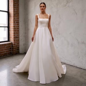 Könnte beinhalten: Ein weißes Brautkleid mit quadratischem Ausschnitt und weitem Rock. Das Kleid hat ein schlichtes Design mit einem eng anliegenden Oberteil und einer langen Schleppe. Das Model steht vor einer Backsteinmauer und einem großen Fenster.
