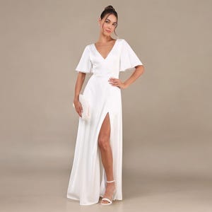 May include: A long, white dress with a V-neck and short, flared sleeves. The dress has a high split on the left leg and is worn with white sandals and a small clutch. The background is a neutral colour. The dress is suitable for special occasions.