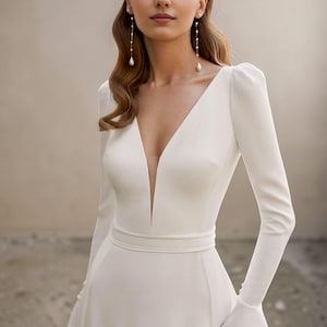 May include: A white bridal gown with a deep V-neck and long sleeves. The dress has a fitted bodice, a defined waist, and a flowing skirt. The sleeves flare slightly at the wrist. The model is wearing long, dangling earrings with pearl drops.