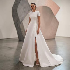 May include: A floor-length white gown featuring a round neckline, short sleeves, and a fitted waistline. The skirt is full with a high front slit that reveals a leg, and the fabric flows into a train. The word Versailles is printed in the corner.